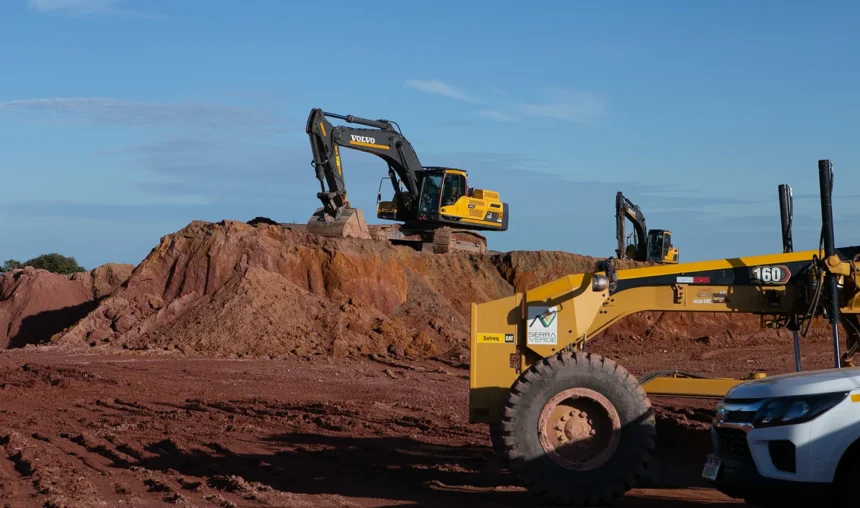 Escavadeiras e equipamentos de construção trabalham em um monte de terra em um canteiro de obras sob um céu azul e limpo, enquanto a equipe avança na extração de terras raras em Serra Verde.