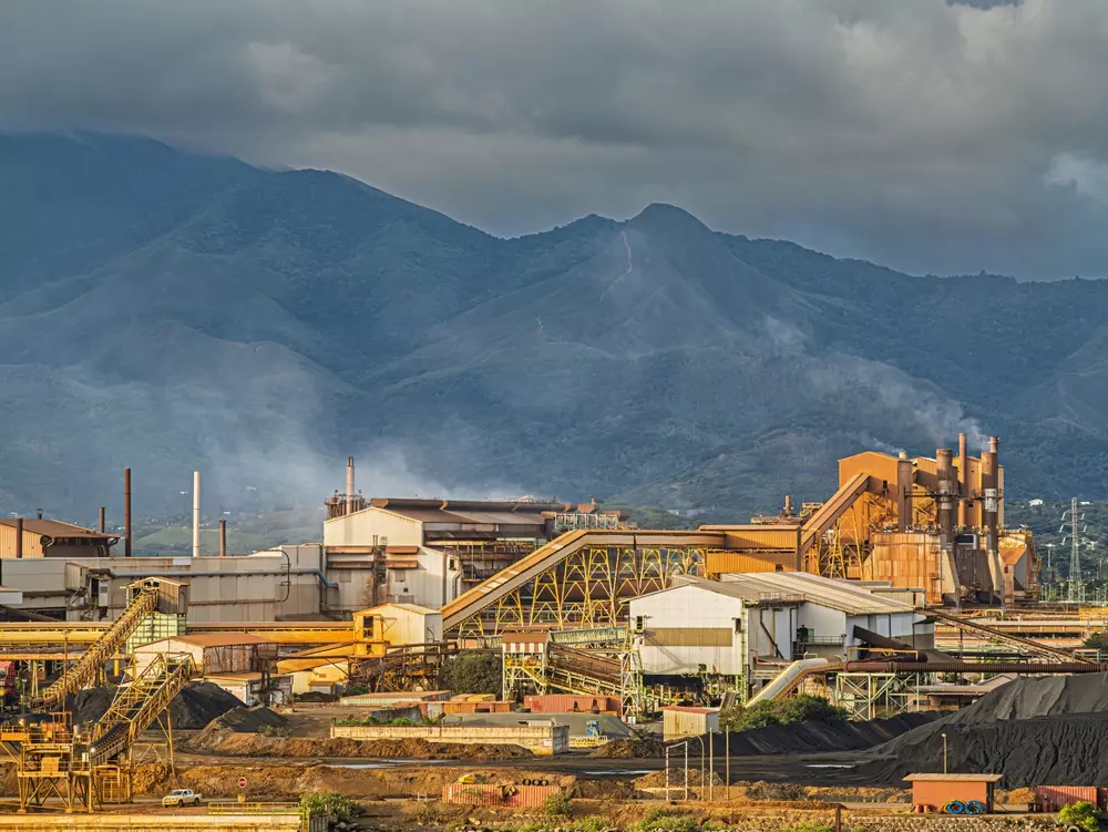 Um complexo industrial especializado em ácido sulfúrico, com chaminés e esteiras transportadoras, fica ao pé de montanhas verdes sob um céu nublado. Pilhas de material preto são visíveis em primeiro plano.