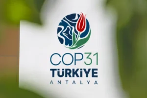 Logotipo da COP31 em Antália, na Turquia, que apresenta um globo estilizado com uma tulipa vermelha e uma folha verde, exibido sobre um fundo branco, parcialmente obscurecido por vegetação desfocada em primeiro plano.