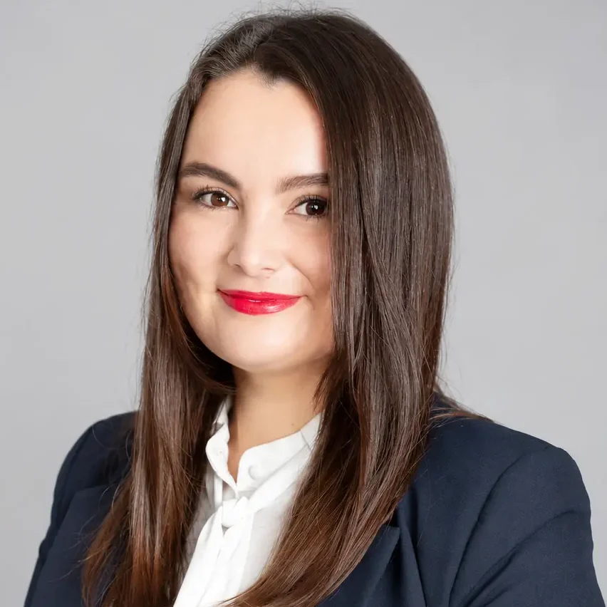 Uma mulher de cabelos castanhos lisos, vestindo um blazer azul-marinho e uma blusa branca, sorri para a câmera diante de um fundo cinza liso.