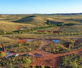 Vista aérea de uma área de mineração com estradas de terra vermelha, máquinas, equipamentos espalhados para a extração de terras raras e vegetação verde cobrindo colinas onduladas sob um céu azul claro.