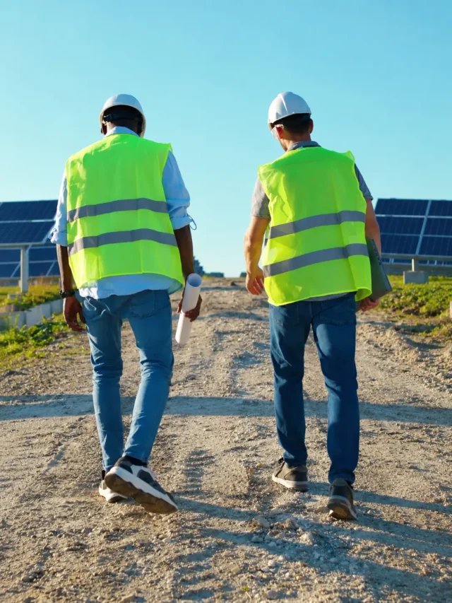 Dois operários da construção civil, vestindo coletes de segurança e capacetes, caminham por um caminho de cascalho em um campo de painéis solares, carregando plantas e um tablet sob um céu azul e limpo.