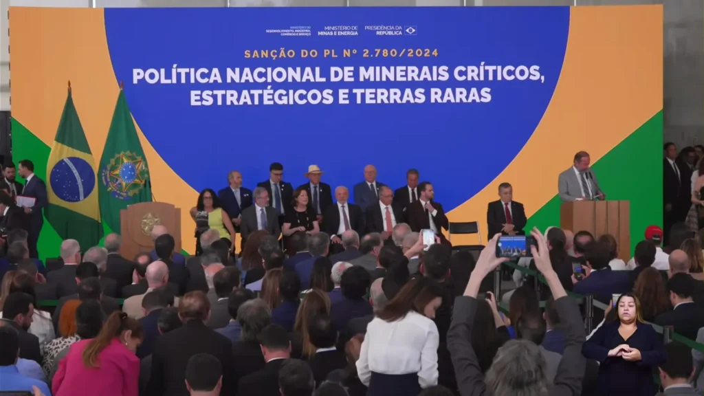 Um grupo de autoridades está sentado e em pé no palco durante um evento sobre a Política Nacional de Minerais Críticos, Estratégicos e de Terras Raras do Brasil (Lei dos Minerais Críticos). Os participantes tiram fotos e há um intérprete de língua de sinais presente.