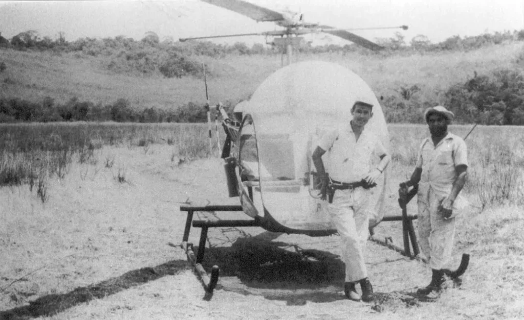 Dois homens est&atilde;o em um campo em Caraj&aacute;s, ao lado de um pequeno helic&oacute;ptero com cabine em forma de bolha.