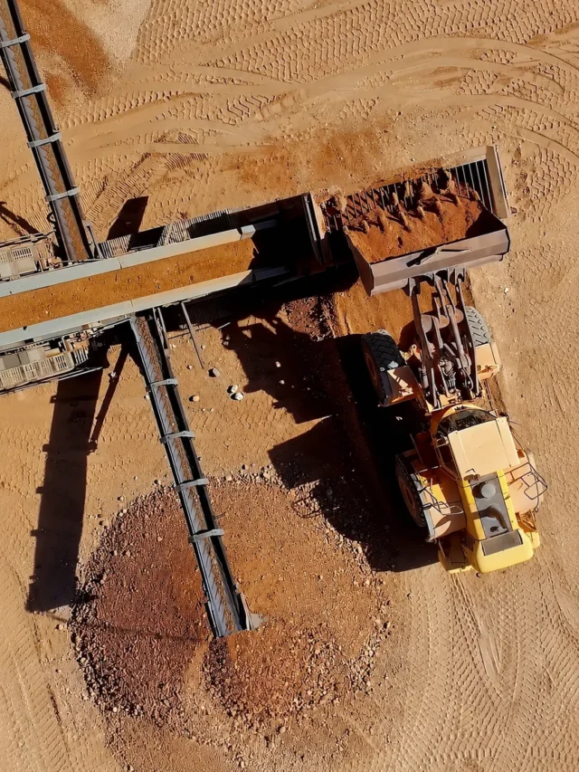 Uma carregadeira frontal amarela despeja terra em um sistema de esteiras transportadoras em um canteiro de obras.